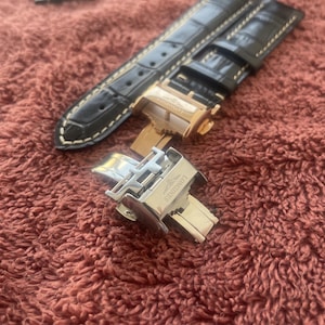 May include: A black leather watch strap with a textured surface and white stitching, paired with a silver-toned metal deployment clasp. The clasp features the engraved Longines brand name on its polished surface, showing a secure locking mechanism.