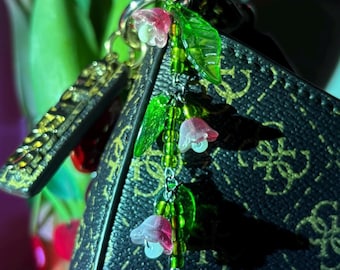 May include: A black handbag with a gold clasp and a floral charm. The charm features pink and white flower beads, green leaf beads, and a gold chain. The bag has a repeating pattern of the letter "G".