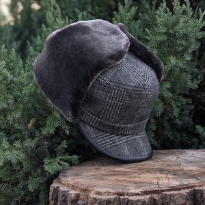 Puede incluir: Un gorro de invierno gris a cuadros con orejeras. El gorro tiene un forro de piel sintética gris oscuro en las orejeras y un borde negro. El gorro está sobre una superficie de madera con follaje verde en el fondo.