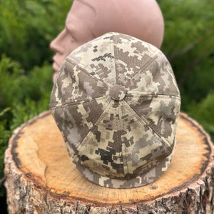 May include: A camouflage newsboy cap in shades of tan, brown, and green. The cap is made of a patterned fabric and features a rounded crown and a short, stiff brim. The cap is displayed on a wooden surface.