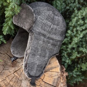 May include: A grey plaid trapper hat with ear flaps and a faux fur lining. The hat has a buckle closure under the chin. The hat is designed to keep the head and ears warm in cold weather. The hat is made of a soft, warm material.