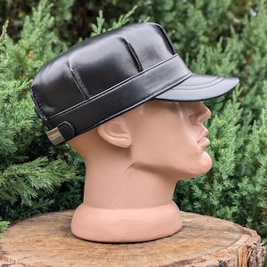Könnte beinhalten: Schwarze Lederkappe mit Visor, präsentiert auf einem Schaufensterkopf. Die Kappe hat ein strukturiertes Design mit einem Band um den Kopf und einem kleinen Metallverschluss. Der Hintergrund zeigt grünes Laub.