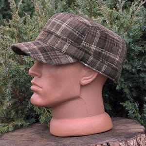 May include: A military-style cadet cap featuring a brown, olive green, and cream plaid pattern. The hat has a flat top, a structured crown, and a short, stiff brim, showcasing a woven fabric texture throughout the entire exterior surface.