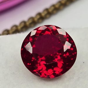 Puede incluir: Una piedra preciosa redonda y facetada de color rojo intenso y vibrante. La piedra presenta un corte brillante con numerosas facetas triangulares y en forma de cometa que reflejan la luz, creando un aspecto brillante y destellante.