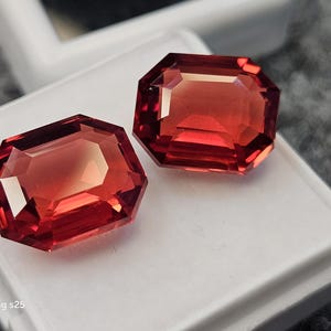 Könnte beinhalten: Zwei lose, rechteckige Edelsteine im Smaragdschliff mit einer leuchtend tiefroten Farbe und facettierten Oberflächen, die das Licht reflektieren. Die Steine liegen auf einem weißen Display-Tablett. Der Text "Samsung s25" ist unten links zu sehen.