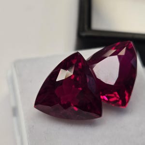 Pode incluir: Duas pedras preciosas triangulares facetadas, de cor vermelha profunda. As gemas são exibidas em uma superfície branca, com uma caixa preta ao fundo. As gemas têm um alto brilho e refletem a luz.