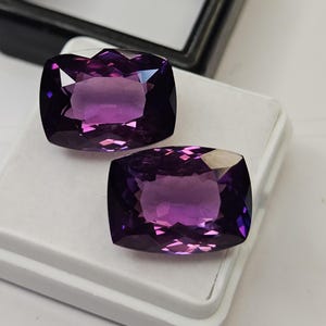 Könnte beinhalten: Zwei kissenförmig geschliffene, tiefviolette Amethyst-Edelsteine mit facettierten Oberflächen. Die Steine sind auf einem weißen, rechteckigen Schmuck-Präsentationskissen platziert. Das Wort surbhit steht in kleinen weißen Buchstaben unten links.