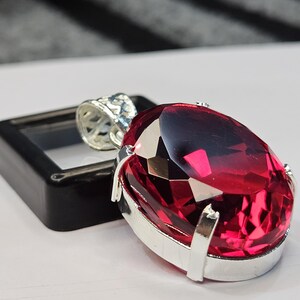 Könnte beinhalten: Ein Anhänger mit einem großen, oval geschliffenen, tiefroten Edelstein in einer silberfarbenen Metallfassung mit Krappen. Das Schmuckstück verfügt über eine dekorative Öse im Filigranstil an der Oberseite und eine facettierte Oberfläche.
