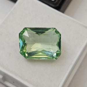 May include: A rectangular, faceted, light green gemstone with a clear, transparent appearance and sharp, angled edges. The stone displays internal light reflections and sits on a white, square display pad with the text surbhit visible.