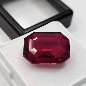 Peut inclure: Une pierre précieuse rouge foncé, taillée en émeraude, avec des surfaces facettées qui reflètent la lumière. La pierre est placée sur un insert en mousse blanche dans une boîte de présentation noire ouverte. Le mot surbhit est écrit en bas à gauche.