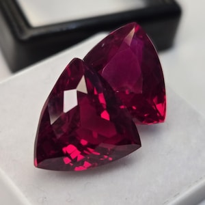 Könnte beinhalten: Zwei lose Edelsteine im Trillion-Schliff mit einer tiefen, leuchtenden magentaroten Farbe. Diese facettierten Steine haben eine dreieckige Form mit spitzen Ecken und mehreren reflektierenden Oberflächen, die das Licht einfangen und klar wirken.