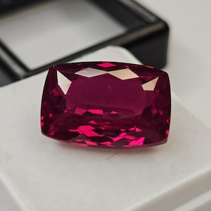 Könnte beinhalten: Ein Edelstein im Kissenschliff mit einer tiefen, leuchtend magentaroten Farbe und facettierter Oberfläche. Der Stein liegt auf einem weißen Schaumstoffeinsatz in einer schwarzen Kunststoffbox. Das Wort surbhit ist unten links aufgedruckt.