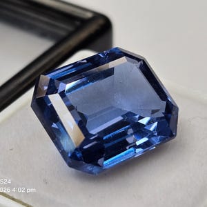 May include: A faceted, rectangular blue gemstone with a clear, transparent appearance and sharp, polished edges. The stone displays deep and light blue tones throughout its structure, reflecting light across its multiple flat, angled surfaces.
