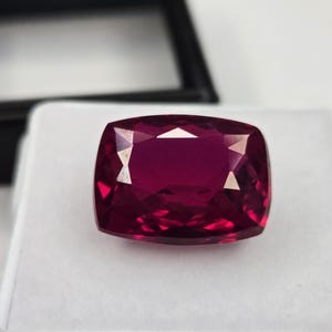 Könnte beinhalten: Ein Edelstein im Kissenschliff mit einer tiefen, gesättigten magentaroten Farbe. Der Stein hat eine facettierte Oberfläche, die das Licht reflektiert und markante geometrische Muster erzeugt. Er liegt auf einem weißen, quadratischen Polster.
