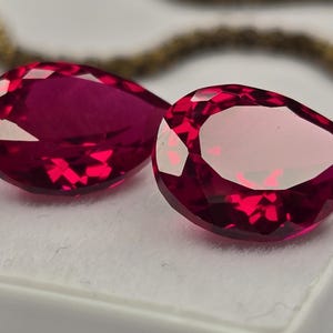 Puede incluir: Dos piedras preciosas de color rojo intenso, talladas en forma de pera con facetas y corte brillante. Las piedras están colocadas una al lado de la otra sobre una superficie blanca, mostrando reflejos internos claros y bordes definidos.