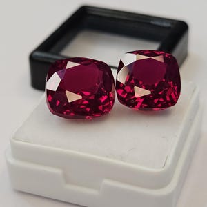 Puede incluir: Dos piedras preciosas de color rojo intenso con talla cojín y superficies facetadas, expuestas sobre un inserto de espuma blanca dentro de una caja de almacenamiento de plástico negro. Las piedras muestran un tono carmesí vivo y saturado.