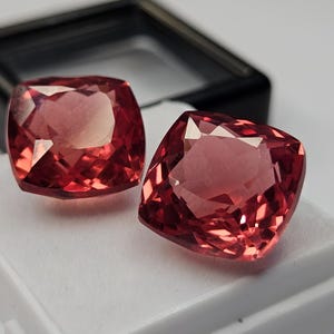Könnte beinhalten: Zwei kissenförmige, rötlich-rosa Edelsteine mit facettierten Oberflächen, die das Licht reflektieren. Die Steine liegen auf einer weißen Schaumstoffeinlage in einer schwarzen Schmuckschatulle. Der Text "surbhit" ist unten links in Weiß gedruckt.