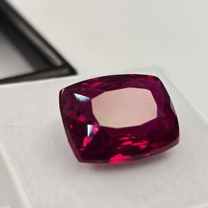 Peut inclure: Une pierre précieuse taille coussin d'une couleur rouge magenta profonde et vibrante avec une surface à facettes qui reflète la lumière. La pierre est placée sur un insert en mousse blanche dans une boîte de présentation noire.