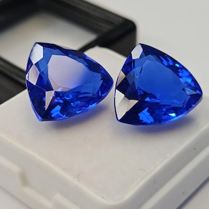Puede incluir: Dos piedras preciosas de color azul vibrante talladas en forma triangular con múltiples facetas que reflejan la luz. Estas piedras sueltas se muestran una al lado de la otra sobre una base de espuma blanca en una caja negra.