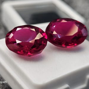 Peut inclure: Deux pierres précieuses rouge foncé de taille ovale avec des surfaces facettées qui reflètent la lumière. Les pierres sont placées côte à côte sur une boîte de présentation rectangulaire blanche avec un couvercle transparent. Le texte Samsung s25 est visible.