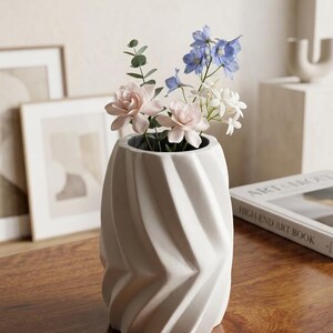 Op de afbeelding: Een witte keramische vaas met een verticale, spiraalvormige geometrische textuur. De vaas bevat een klein boeket van lichtroze bloemen, kleine witte bloesems en blauwe ridderspoorstelen met groene eucalyptusbladeren in de opening.