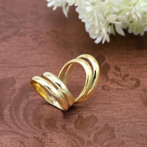 Può includere: Un anello color oro con tre fasce collegate. Le fasce sono lisce e curve, creando un design unico. L'anello è esposto su una superficie marrone con un motivo floreale, con fiori bianchi sullo sfondo.