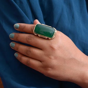 Op de afbeelding: Een grote, rechthoekige ring met een groene edelsteen in een goudkleurige, sierlijke zetting. De ring wordt gedragen op een hand met turquoise nagellak, tegen een blauwe achtergrond.