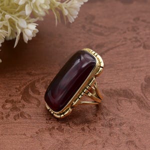 Könnte beinhalten: Goldfarbener Ring mit einem großen, rechteckigen, tief burgunderfarbenen Stein. Der Ring hat eine detaillierte, strukturierte Fassung und ein geteiltes Schaftdesign. Der Ring befindet sich vor einem braunen Hintergrund mit floralen Mustern.