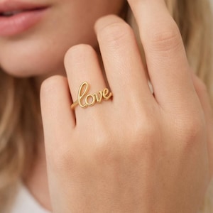Può includere: Un anello dorato con la parola "love" scritta in corsivo. L'anello è indossato su un dito, con la parola "love" ben visibile. Il design dell'anello è semplice ed elegante, adatto all'uso quotidiano.