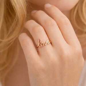 Puede incluir: Un anillo dorado con la palabra "love" escrita en cursiva. El anillo se lleva en un dedo, con la mano y parte del brazo visibles. El anillo es el punto focal, con un fondo suave.