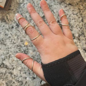 Puede incluir: Una mano que lleva varias férulas para dedos de color plateado. Las férulas son curvas y están diseñadas para adaptarse a los dedos, proporcionando soporte. La mano descansa sobre una superficie, siendo las férulas el foco principal de la imagen.