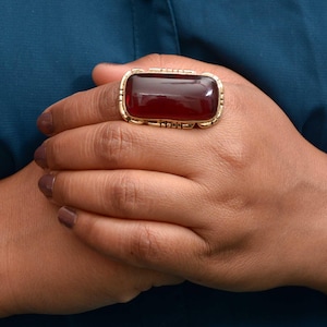 Puede incluir: Un anillo dorado con una piedra grande, rectangular y de color rojo intenso. El anillo tiene un engaste ornamentado y detallado alrededor de la piedra. El anillo se muestra en una mano con esmalte de uñas oscuro, sobre un fondo verde azulado.