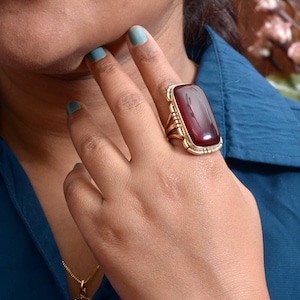 Op de afbeelding: Een grote, rechthoekige ring met een dieprode steen gezet in een goudkleurig metalen frame. De ring heeft een gedetailleerd ontwerp aan de zijkanten en wordt aan een vinger gedragen. De achtergrond toont een blauw overhemd en een ketting.