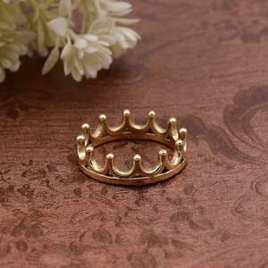 Può includere: Un anello a corona color oro con un design dettagliato. L'anello presenta una serie di archi rivolti verso l'alto, ciascuno sormontato da una piccola perla arrotondata. L'anello è posto su uno sfondo marrone con un motivo floreale e fiori bianchi.