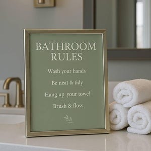 Puede incluir: Un letrero enmarcado con el texto "BATHROOM RULES" en un baño. Las reglas se enumeran debajo del título. Toallas blancas enrolladas están apiladas en la encimera. Un grifo dorado y una lámpara de pared son visibles.