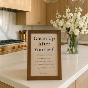 May include: A framed sign with the text "Clean Up After Yourself" in a kitchen setting. The sign includes bullet points with cleaning instructions. The kitchen features light wood cabinets, a white countertop, and a vase of white flowers.