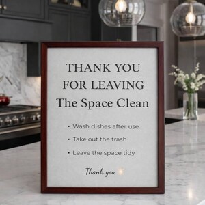 May include: A framed sign with a dark wood border displays the text "THANK YOU FOR LEAVING The Space Clean." Instructions include washing dishes, taking out the trash, and leaving the space tidy. The sign is on a marble countertop.