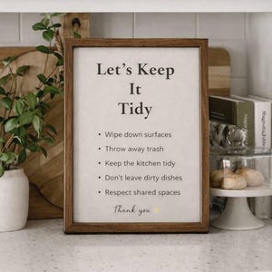 以下が含まれることがあります： 「Let's Keep It Tidy」の文字と掃除のリマインダーが書かれた額入りのサイン。茶色の木製フレームに入っており、白いカウンタートップに置かれています。テキストは黒色です。