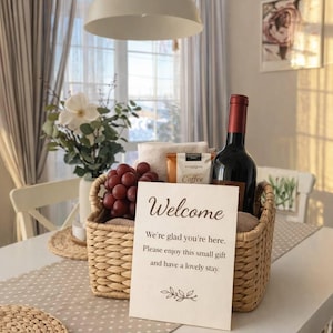 Puede incluir: Una cesta de mimbre llena de una botella de vino tinto, uvas, café y una toalla doblada. Una tarjeta con la palabra "Bienvenida" está frente a la cesta. El texto de la tarjeta dice: "Nos alegra que estés aquí. Disfruta de este pequeño regalo y que tengas una estancia agradable."