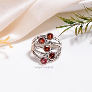 Può includere: Un anello d'argento con sei pietre preziose rotonde, rosso intenso. L'anello ha un design a fascia strutturata e il nome del marchio "Risergemsandjewel" è stampato in basso.