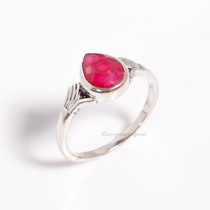 May include: A silver ring featuring a pear-shaped, vibrant red gemstone. The ring has a detailed, leaf-like design on either side of the gemstone. The ring is set against a white background, highlighting the jewelry's features.