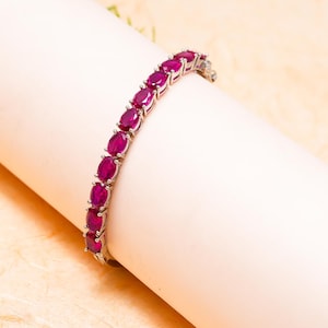 May include: A silver-toned bracelet featuring a series of oval-cut, vibrant magenta-red gemstones set in individual metal prongs. The bracelet follows a curved line, showcasing a consistent row of faceted stones that catch the light.