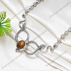 Könnte beinhalten: Eine silberfarbene Halskette mit Schlangenmotiv. Die Halskette hat einen zentralen, ovalen, braunen Edelstein. Der Schlangenkörper ist mit strukturierten Mustern versehen. Die Halskette wird auf einem weißen, seidigen Stoff präsentiert.