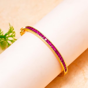 Puede incluir: Una pulsera rígida articulada de tono dorado que presenta una hilera continua de piedras de corte cuadrado en color rosa magenta vibrante, engastadas en canal. La pulsera tiene un acabado metálico pulido y un cierre visible en el lateral.