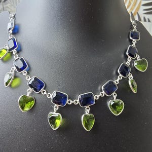 May include: A silver necklace featuring a series of dark blue, rectangular gemstones interspersed with heart-shaped, light green gemstones. The necklace is displayed on a black mannequin.