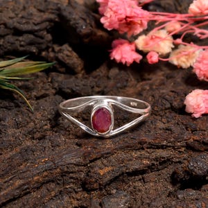 May include: A silver ring with a ruby gemstone. The ring features a split band design that curves around the oval-shaped, deep red ruby. The ring is set against a dark, textured background with pink flowers.