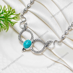 以下が含まれることがあります： 中央にターコイズ色の楕円形の石をあしらったシルバーカラーのネックレス。デザインは、テクスチャ加工が施された2つのヘビのモチーフを取り入れています。チェーンは小さく丸みを帯びたビーズで構成されており、ユニークで人目を引くアクセサリーです。