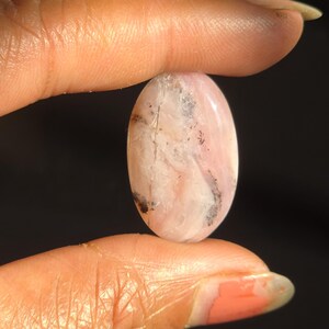 May include: Oval-shaped pink gemstone with white and black inclusions. The polished stone is held between two fingers against a dark background. The gemstone has a smooth, glossy surface and a natural, organic appearance.