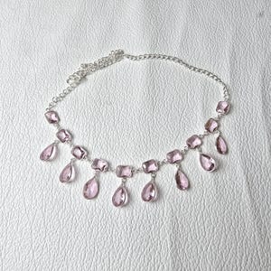 May include: A silver-toned necklace featuring rectangular and teardrop-shaped pink gemstones. The necklace has a delicate chain and is displayed on a white surface. The gemstones are evenly spaced along the necklace.