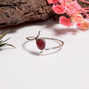 May include: A silver ring with a large, oval, deep red gemstone. The ring has a unique design with curved, stylized elements. The ring is set on a white surface, with pink flowers and a piece of wood in the background. The gemstone is approximately 1.5 cm in length.